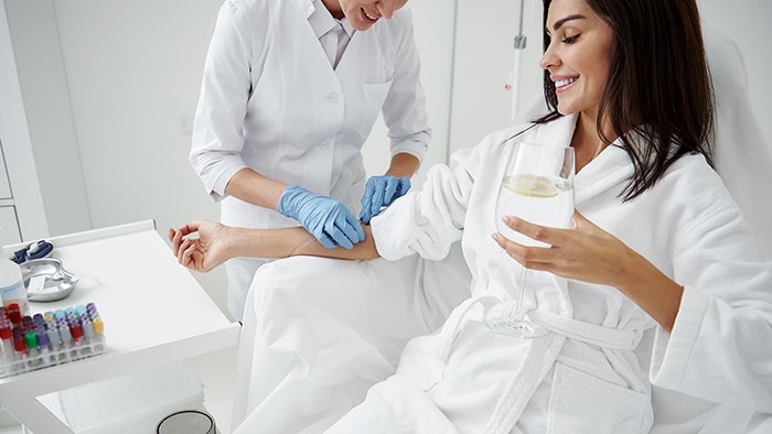 Cropped portrait of beautiful woman in white bathrobe sitting in armchair and receiving IV infusion. She is holding glass of beverage with lemon and smiling